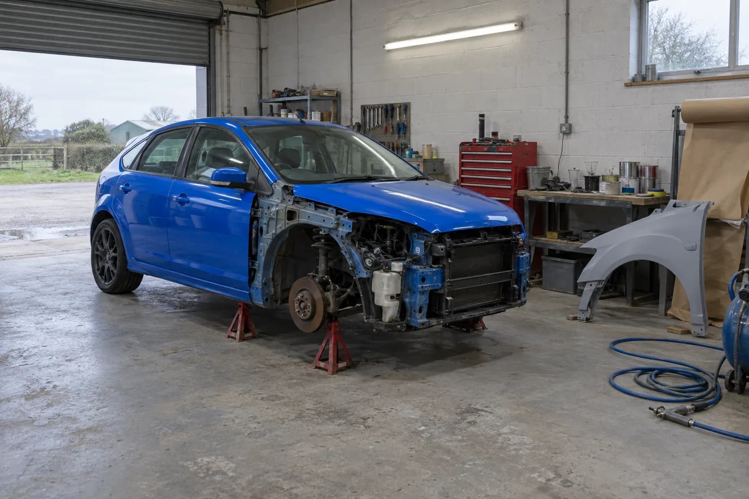Illustration: a blue hatchback on axle stands in the workshop with its front wing off, the replacement wing waiting on the bench
